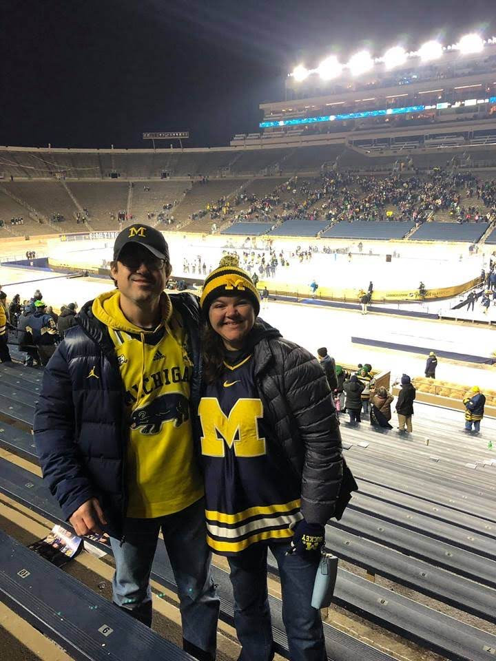A picture of Bernadette Skodack and her husband wearing University of Michigan clothing. They are smiling at the camera, with an ice rink behind them.